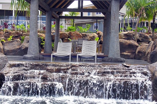 Two beach chairs sitting in the water with a waterfall right in front of them. Lava rack surrounds the pool.