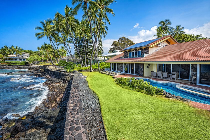Backyard with seawall and green grass. House and pool with beach chairs and palm trees.